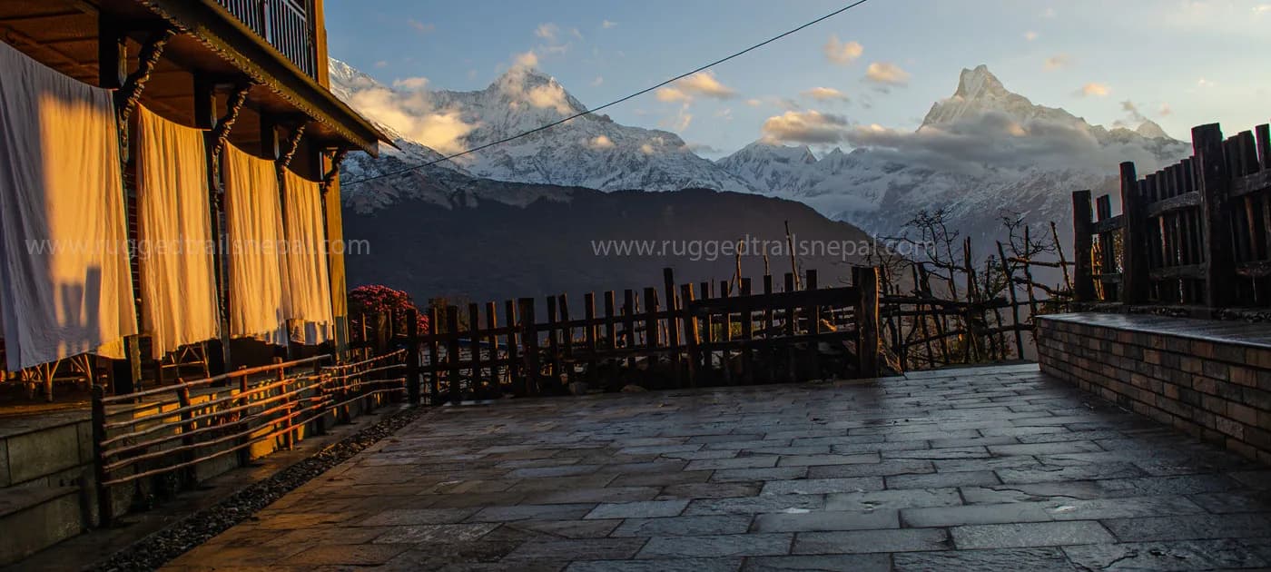 A stone patio and traditional lodge balcony illuminated by warm golden sunlight, looking out toward the snow-capped peak of Machhapuchhre (Fishtail) and the Annapurna range