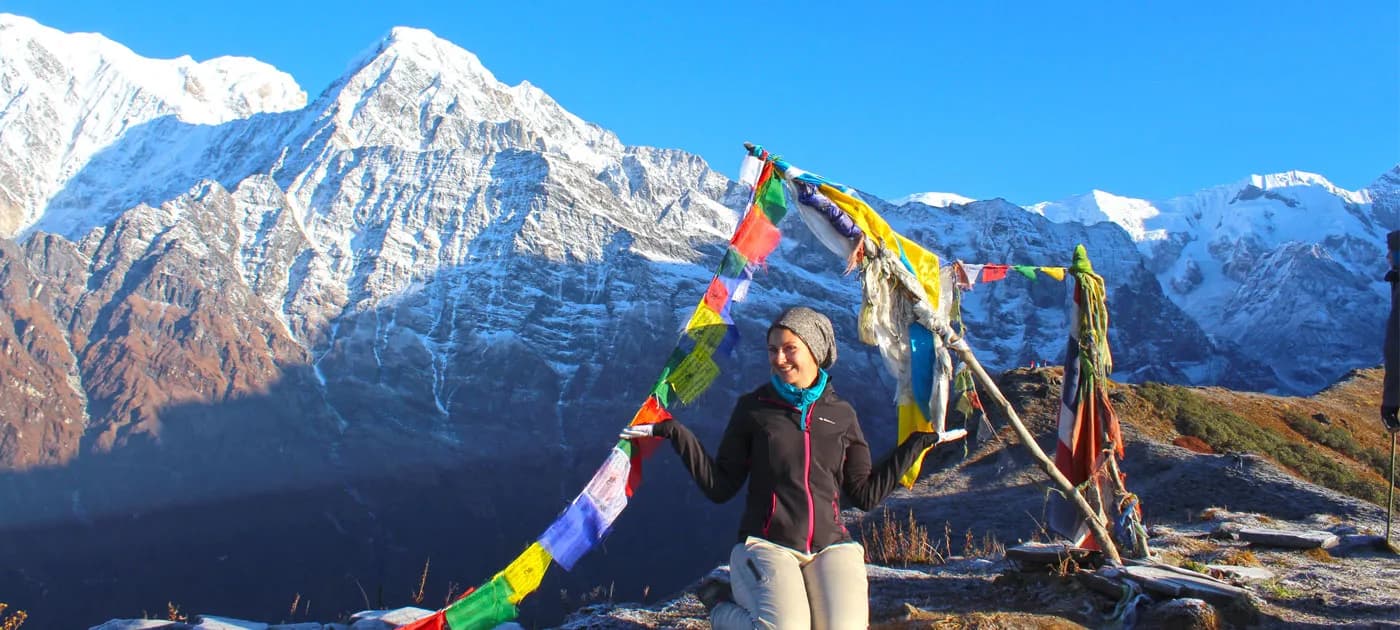 A female trekker posing under colorful Buddhist prayer flags framed against snow-capped mountains on a short trek near Pokhara.