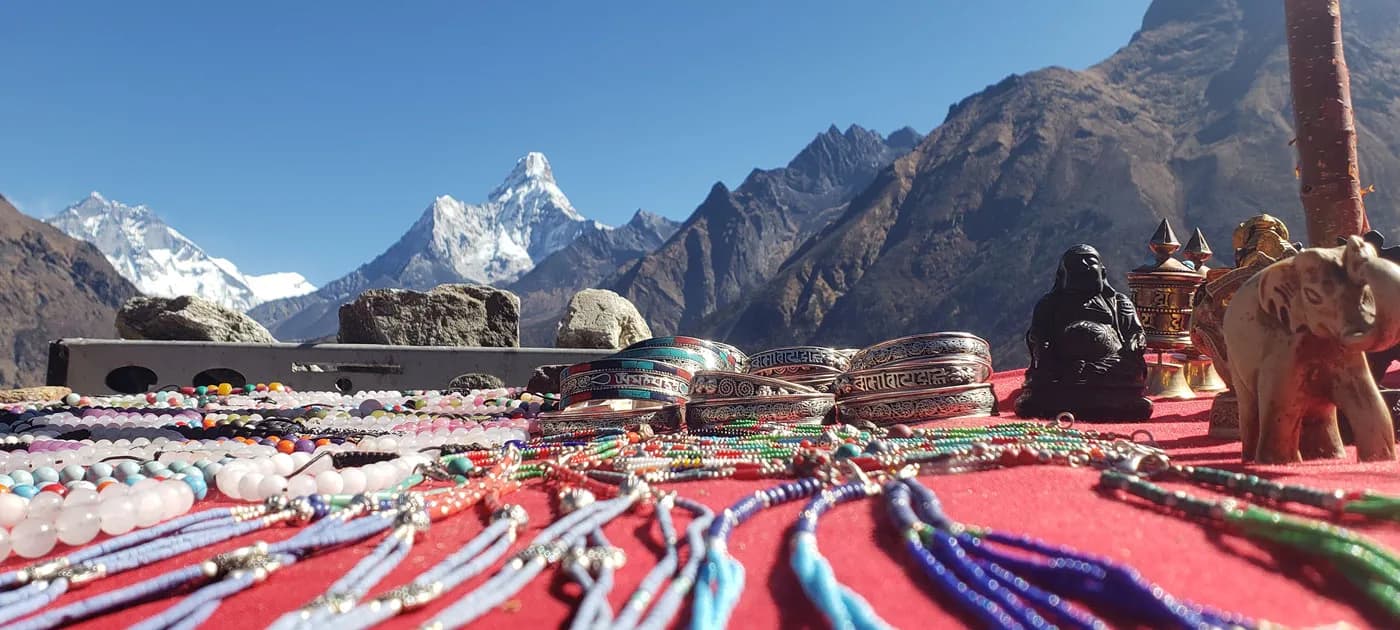 A vibrant table display of bead bracelets, Buddha statues, and souvenirs set against a clear blue sky and snow-capped Himalayan peaks including Ama Dablam