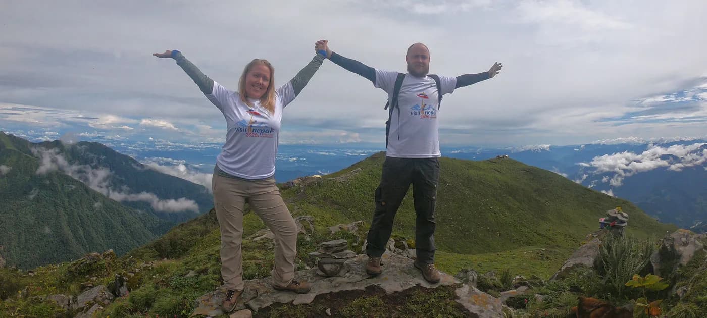 Two hikers in matching white promotional t-shirts celebrating on a rocky ridge with panoramic views of lush green valleys and cloud-filled hills.