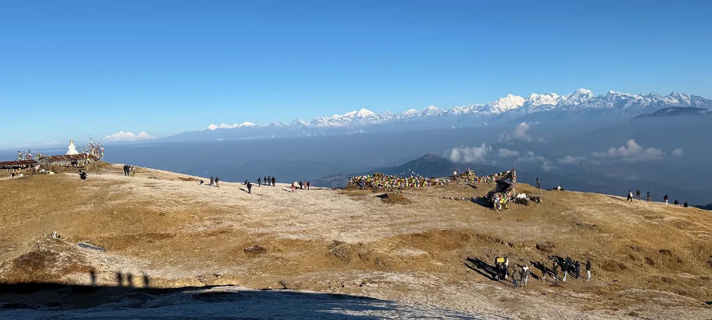 A vast high-altitude grassy plateau at Sailung in Dolakha with scattered visitors, prayer flags, and a distant Himalayan mountain panorama under a clear blue sky.