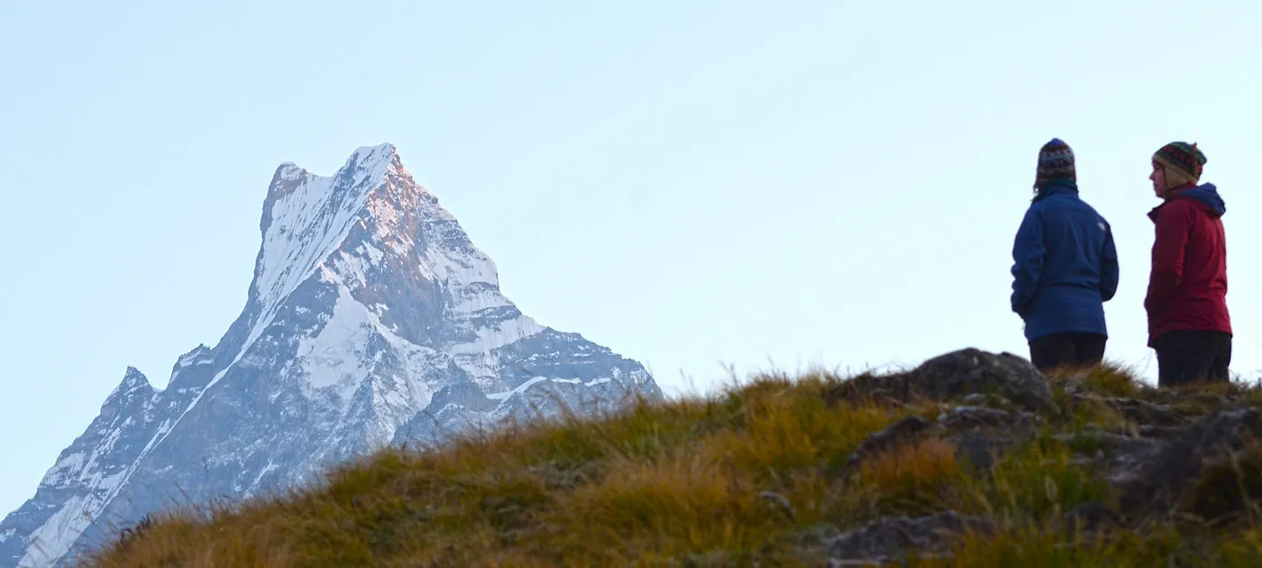 Two hikers standing on a grassy ridge looking out at the iconic fishtail peak of Mount Machhapuchhre in the Annapurna region.