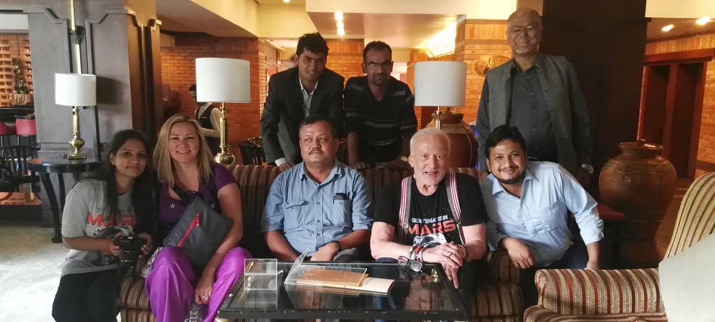 Buzz Aldrin wearing a Destination Mars t-shirt seated on a couch posing for a photo with a group of local hosts in Nepal.