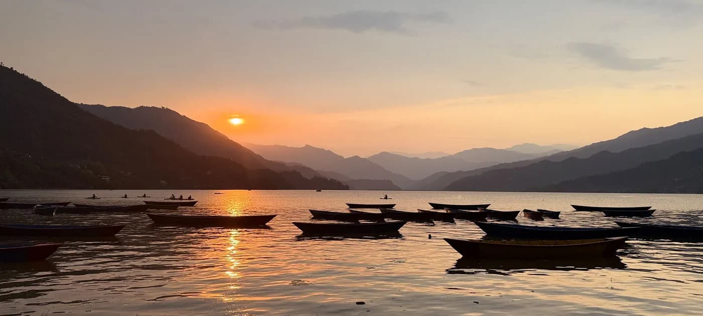 Silhouetted wooden boats resting on Phewa Lake as the sun sets behind misty mountain ridges, casting a warm golden glow across the water