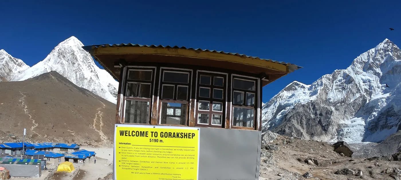Yellow and white welcome sign board at Gorakshep listing altitude information and trail details in front of snowy Himalayan mountains.
