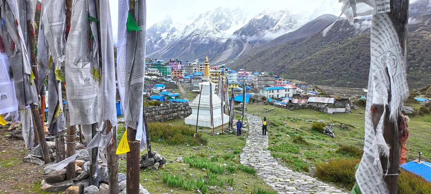 A stone pathway framed by vertical prayer flag poles leading toward a white Buddhist chorten and a mountain village in Kyanjin Gompa