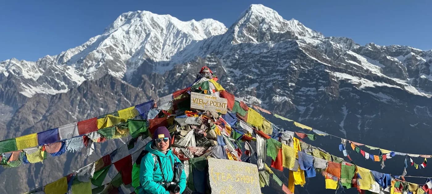 A female trekker smiling in front of a prayer flag monument marked "VIEW POINT" with snow-capped Annapurna peaks behind her.