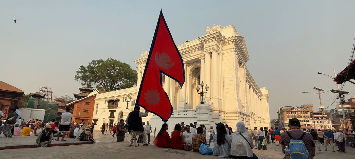 Locals and tourists sitting near the white neoclassical Gaddi Baithak palace in Kathmandu Durbar Square with a large Nepalese national flag in the foreground.