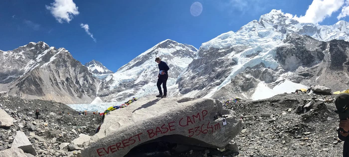 A trekker standing on top of the large boulder painted with EVEREST BASE CAMP 5364m against massive snow-capped Himalayan peaks