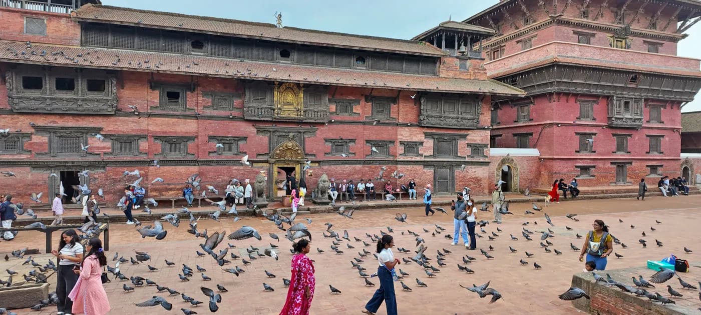 People walking and pigeons flying in front of historic brick palaces at Patan Durbar Square