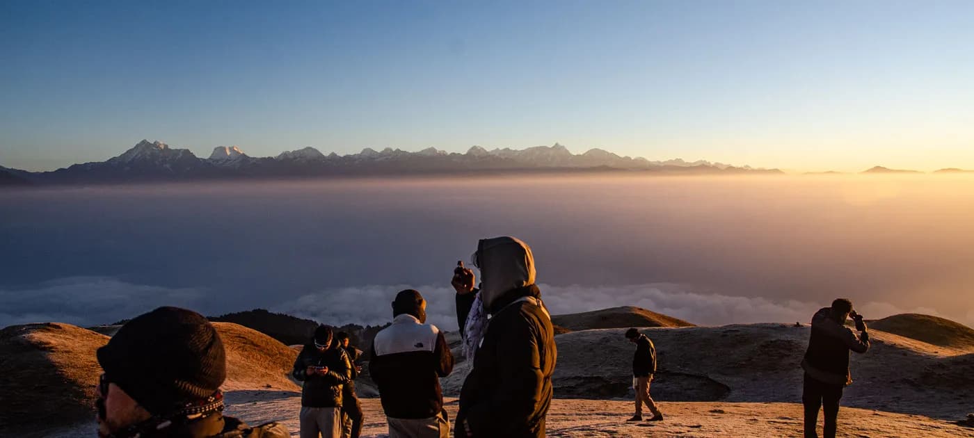 Group of travelers bundled in warm jackets watching a stunning golden sunrise above the cloud line with mountain peaks in the distance.