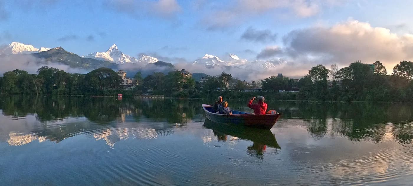 A wooden boat with passengers floating on tranquil Phewa Lake at sunrise with misty mountain reflections in the water.