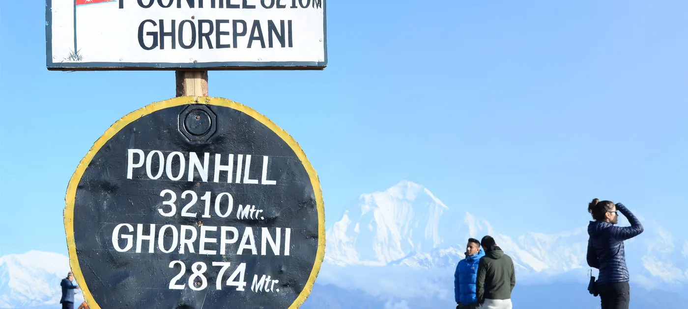 A close-up view of the circular Poon Hill 3210m elevation signpost with visitors admiring distant snow-capped mountains.