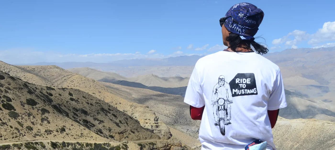 Traveler wearing a 'Ride to Mustang' t-shirt looking out over the mountain valley landscape in Mustang, Nepal.