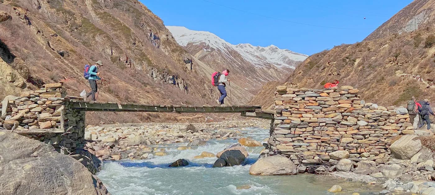 Trekkers crossing a rustic wooden bridge supported by stacked stone piers over a rushing mountain river with snow-draped Himalayan mountains in the background