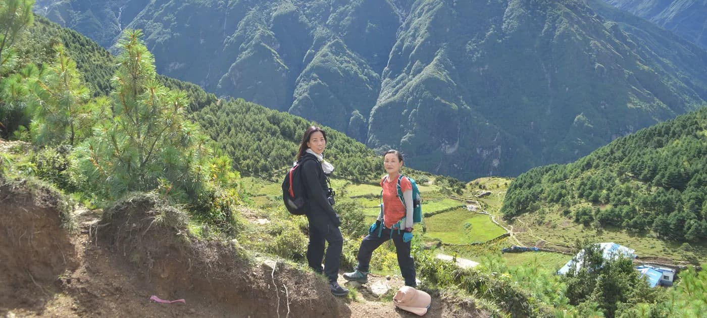 Two female trekkers carrying backpacks standing on a scenic mountain trail overlooking deep green valleys in the Everest region