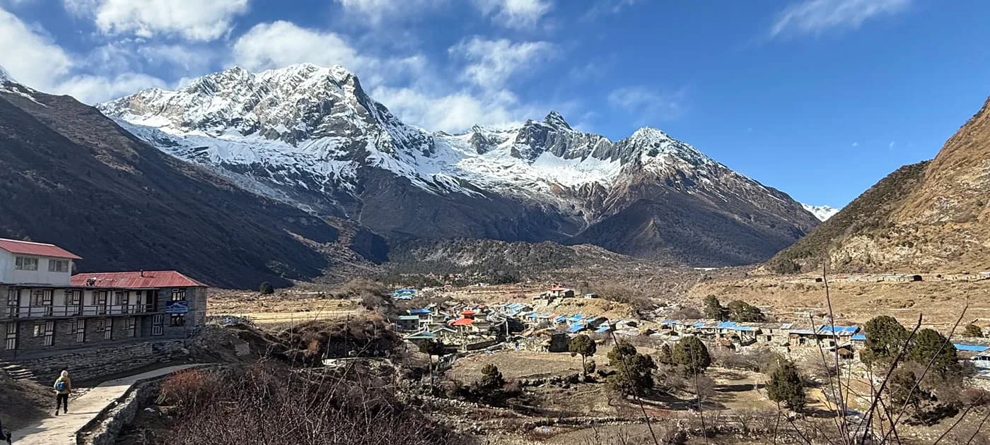 A wide landscape view of Samagaun village with colorful teahouse roofs and stone lodges situated in an alpine valley beneath majestic snow-covered Himalayan peaks.