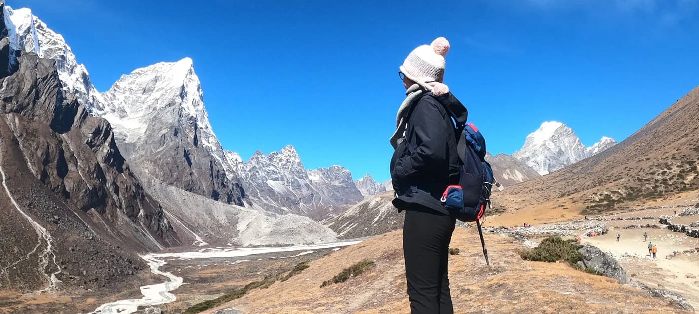 A female hiker with a backpack standing on a ridge looking out over a wide Himalayan valley and steep sharp mountain peaks