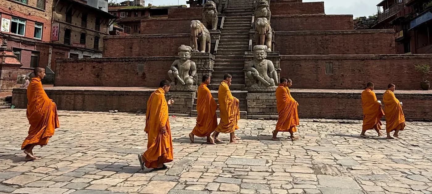 Buddhist monks in bright orange robes walking single file across the stone plaza past guardian statues of Nyatapola Temple in Bhaktapur