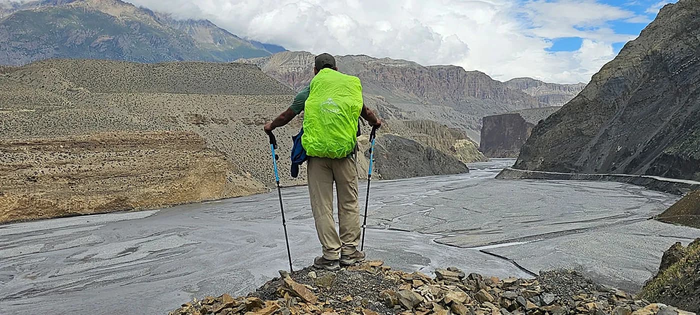 Rear view of a trekker with trekking poles wearing a bright green rain cover over his backpack, looking out over a river valley.