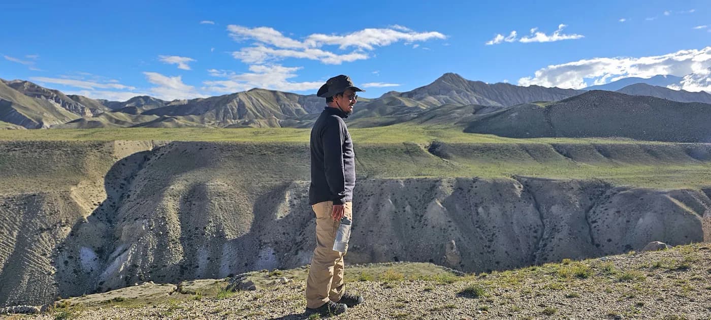 Trekker in a wide-brimmed sun hat standing on a cliff edge looking across the vast eroded canyons and desert plateau of Upper Mustang.
