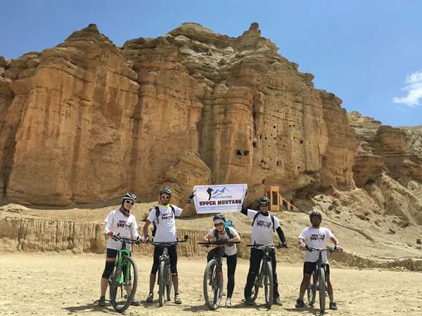 A group of mountain bikers wearing helmets, matching shirts, and sunglasses standing with their bikes holding an Upper Mustang banner in front of high, eroded sandstone cliffs