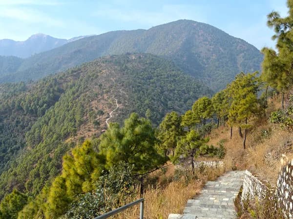 A paved stone walking path with a metal handrail winding through pine trees along a mountain ridge leading towards Champadevi Hill