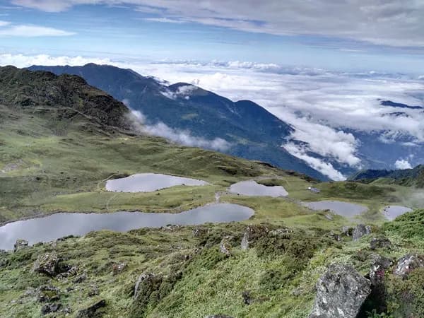 High-angle aerial landscape view of the five sacred glacial lakes of Panch Pokhari spread across green alpine pastures with surrounding mountain ridges and clouds below