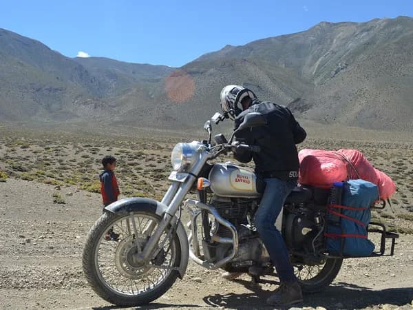 A motorcyclist wearing a helmet and riding gear seated on a classic silver Royal Enfield loaded with luggage along a dirt road in a dry, mountainous landscape