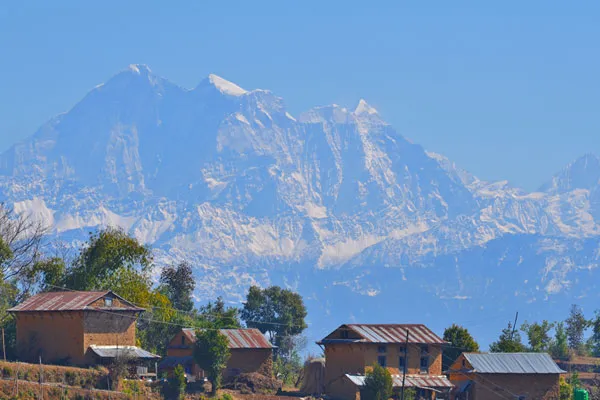 Traditional clay and brick village homes set against a clear blue sky and towering snow-covered Himalayan peaks.