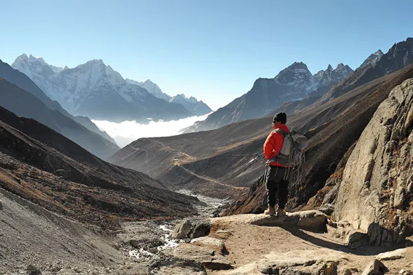 A trekker in an orange jacket with a backpack standing on a high-altitude rocky trail looking down into a mist-filled mountain valley in the Everest region.