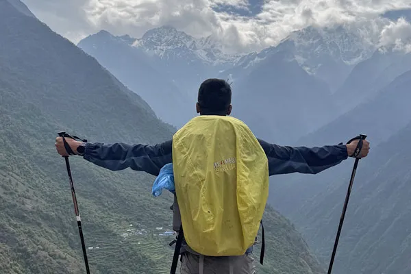 A male trekker with trekking poles and a yellow backpack cover enjoying views of snow-capped mountains and deep valleys along a short Nepal trekking route.