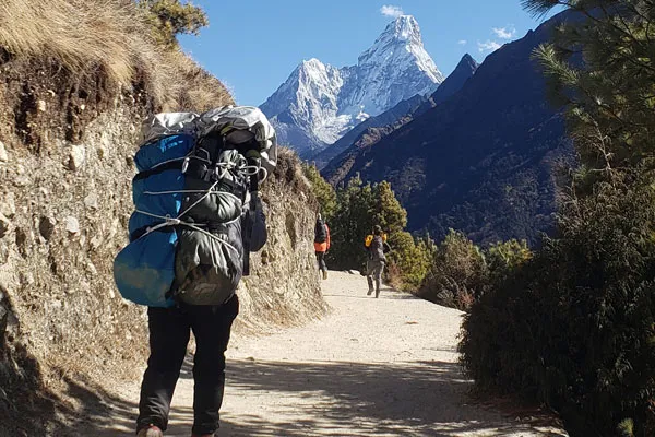 A local porter carrying a large bundle of trekker gear along a wide mountain path with Ama Dablam towering in the background