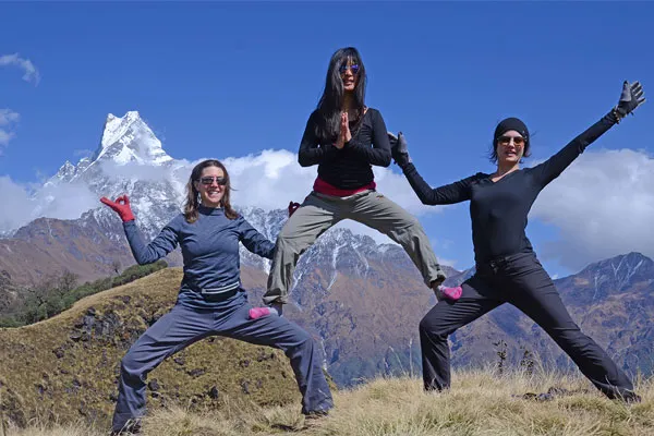 Three female practitioners doing a fun acro-yoga group pose on a grassy ridge with the distinctive peak of Mount Machhapuchhre behind them