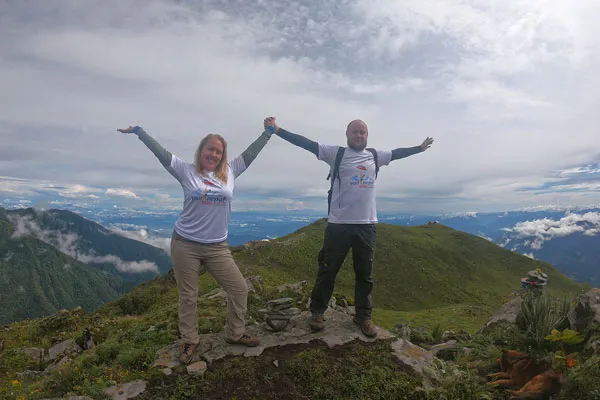 A man and woman wearing matching white "Visit Nepal 2020" t-shirts standing on a mountain peak with arms raised high against a valley backdrop.