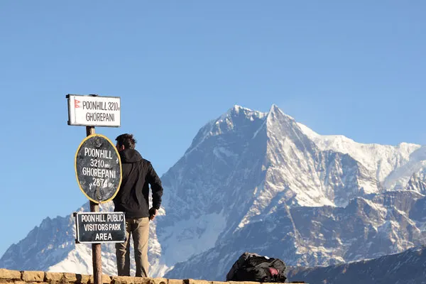A trekker standing next to the Poon Hill 3210m elevation signpost looking out at Mount Machhapuchhre in the Annapurna range