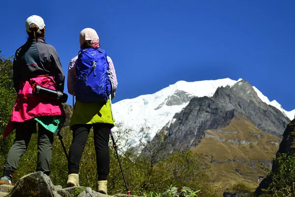 Two female trekkers carrying backpacks standing on a stone path and looking toward a snow-capped Himalayan mountain peak
