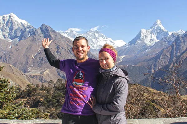 A smiling man in a purple t-shirt and a woman in a grey jacket standing together on a vantage point with Mount Everest
