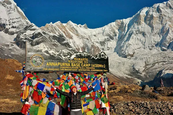 A female trekker posing with arms outstretched in front of the Annapurna Base Camp signpost decorated with colorful prayer flags under a clear blue sky