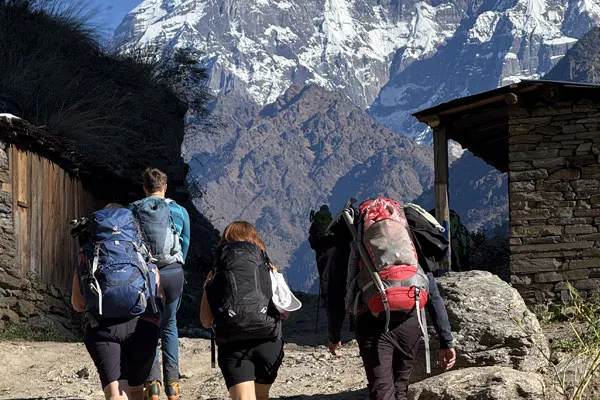 A group of trekkers wearing heavy backpacks walking up a gravel path past traditional stone houses toward towering snow-capped mountains