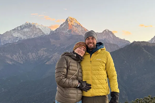 A smiling couple wearing warm jackets and beanies posing together on a viewpoint against the golden sunrise over the Annapurna mountain range