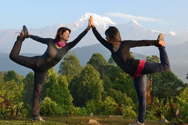 Two female yogis holding partner dancer poses touching hands outdoors with lush green hills and snow-capped Annapurna peaks in the background