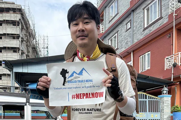 A male trekker holding a Rugged Trails Nepal printed sign with the hashtag NEPALNOW in front of a residential building in Kathmandu