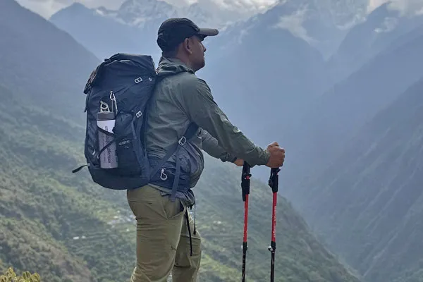 A male trekker in a green jacket and cap holding trekking poles and carrying a blue backpack while looking across a misty mountain valley