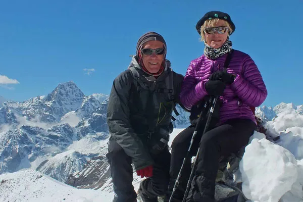 A man and woman standing together next to colorful Buddhist prayer flags on Kala Patthar viewpoint with snow-covered Himalayan peaks in the background.