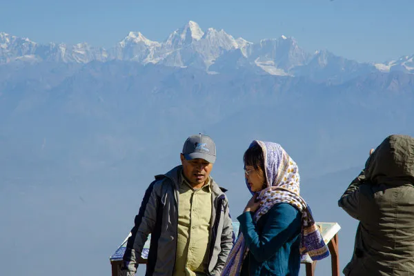 A local guide in a cap explaining points of interest to a tourist at a hill viewpoint with distant snow-capped mountains in the background