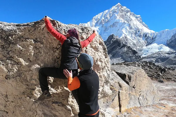 A female hiker bouldering on a large rock supported by a spotter with high-altitude snow peaks including Nuptse in the background.