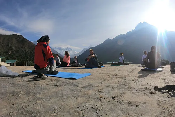 A group of people sitting on blue yoga mats outdoors practicing meditation under bright sunlight against a mountain backdrop in Everest
