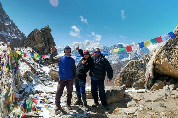 Three trekkers wearing winter gear and sunglasses posing cheerfully together on a high mountain pass adorned with colorful prayer flags