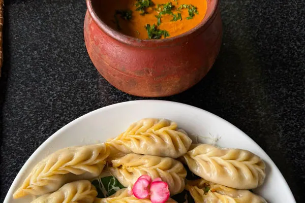 A white plate filled with steamed Nepalese momo dumplings served alongside a clay pot of savory orange dipping chutney